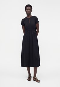 Femme portant une robe midi noire à manches courtes avec une taille froncée et une fente à l'avant, associée à des sandales noires avec des accents dorés.