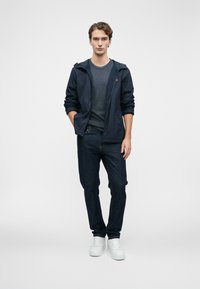 Νεαρός άνδρας που στέκεται μπροστά σε απλή φόντο, φορώντας ναυτική κουκούλα, σκούρο γκρι μπλουζάκι, σκούρα τζιν και λευκά αθλητικά παπούτσια.