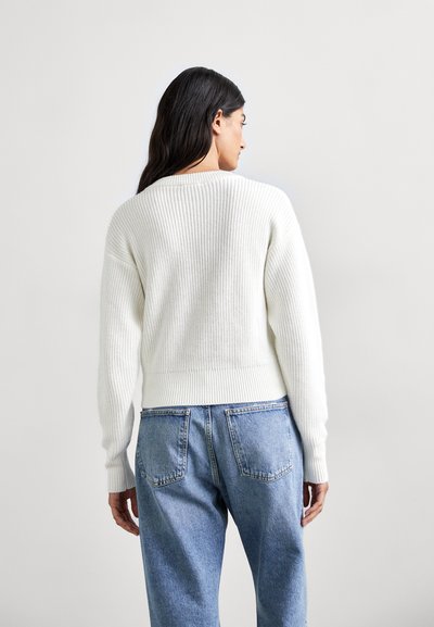 Witte gebreide trui met ribstructuur, cropped pasvorm en lange mouwen, gecombineerd met blauwe spijkerbroek, gezien vanaf de achterkant.