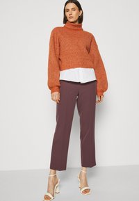 Rostorange chunky stickad turtlenecktröja lagd över en vit skjorta, parat med högmidjade plommonfärgade byxor och vita remsandaler.
