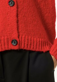 Roter gestrickter Cardigan mit geripptem Saum und Bündchen, ausgestattet mit einer schwarzen Knopfverschluss. Strukturiert mit sichtbaren, flauschigen Strickdetails.