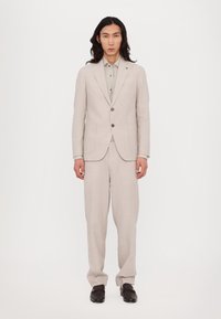 Costume en lin beige clair avec épaules structurées, blazer à un bouton et pantalon large. Associé à une chemise à col gris clair.