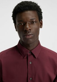 Burgundy button-up shirt with a collar, featuring a smooth texture and shiny black buttons on a solid background.