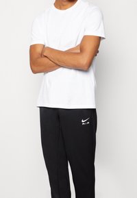 Bílá tričko s kulatým výstřihem spárované s černými tepláky s bílým logem Nike Air. Tričko má krátké rukávy a standardní střih.