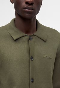 Gros plan d'une personne portant un cardigan boutonné vert olive avec un col et une petite étiquette Calvin Klein sur la poitrine.