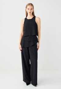 Schwarzes ärmelloses Top mit gestaffeltem Design, kombiniert mit weit geschnittenen schwarzen Hosen. Glatter, matter Stoff mit leichtem Glanz.