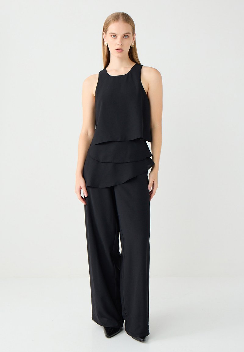 Schwarzes ärmelloses Top mit gestaffeltem Design, kombiniert mit weit geschnittenen schwarzen Hosen. Glatter, matter Stoff mit leichtem Glanz.