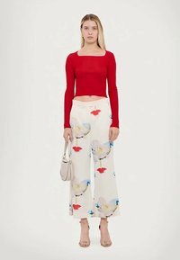 Femme portant un crop top rouge à manches longues, un pantalon blanc à jambes larges avec imprimé floral, des talons beiges, et tenant un petit sac à main beige.
