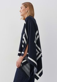 Navy Poncho aus weichem Material mit geometrischen weißen Streifen, offenen Seiten und dreieckiger Form. Eignet sich für verschiedene Outfits.