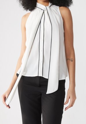 Mujer con blusa blanca sin mangas con ribetes negros y detalle de bufanda larga, combinada con pantalones negros, de pie frente a un fondo blanco.