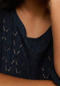 Gros plan sur la partie supérieure de la poitrine et le bras d'une personne portant un haut tricoté bleu foncé avec un motif délicat en dentelle.