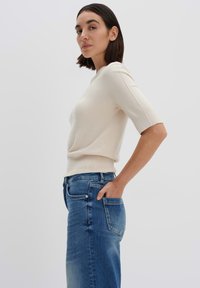 Hellbeige kurzärmliger Pullover mit tailliertem Schnitt; kombiniert mit hochgeschnittenen blauen Jeans. Dezente Struktur auf dem Stoff sichtbar.