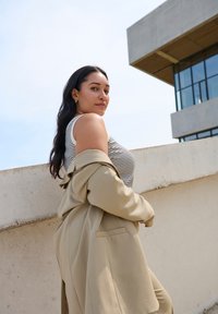 Blazer beige oversize sur un débardeur rayé, avec un col rond. Texture lisse, cheveux longs et foncés, avec des bijoux minimum.