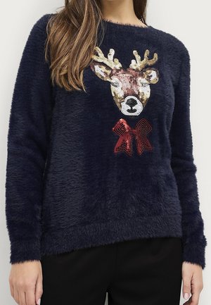 Femme portant un pull en maille peluche bleu marine avec un visage de renne en sequins et un nœud rouge sur le devant.