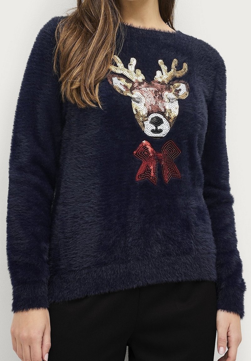 Femme portant un pull en maille peluche bleu marine avec un visage de renne en sequins et un nœud rouge sur le devant.