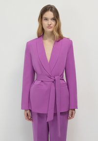 Giacca su misura viola con scollo a V profondo, maniche lunghe e cintura anteriore. Presenta due tasche laterali e una texture liscia.