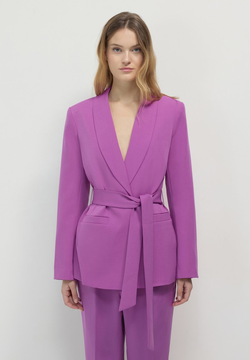 Giacca su misura viola con scollo a V profondo, maniche lunghe e cintura anteriore. Presenta due tasche laterali e una texture liscia.