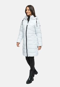 Mujer vestida con un abrigo de invierno acolchado con capucha en gris claro, pantalones negros y botas negras hasta el tobillo, de pie con una ligera sonrisa.