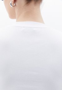 Bílý pruhovaný top s kulatým výstřihem, vyrobený z texturované tkaniny a s minimálními švy na zadní straně a výstřihu. Viditelné stříbrné kruhové náušnice.