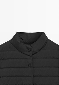 Giacca imbottita nera con collo alto, caratterizzata da una sottile cucitura orizzontale e due chiusure con bottoni al collo. Tessuto liscio e leggero.