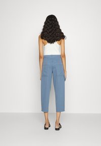 Nainen, jolla on aaltoilevat tummat hiukset, valkoinen hihaton toppi, vaaleansiniset, leveälahkeiset ja lyhennetyt housut suurilla takataskuilla sekä mustat slip-on-kengät.