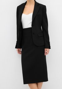 Femme portant un blazer noir sur mesure et une jupe assortie longueur genoux par-dessus un haut blanc ajusté, debout devant un fond blanc uni.
