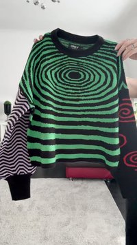 Zielono-czarny sweter we wzory z spiralnymi motywami i paskami. Lewy rękaw ma naprzemienne czarno-białe wzory w zygzaki.