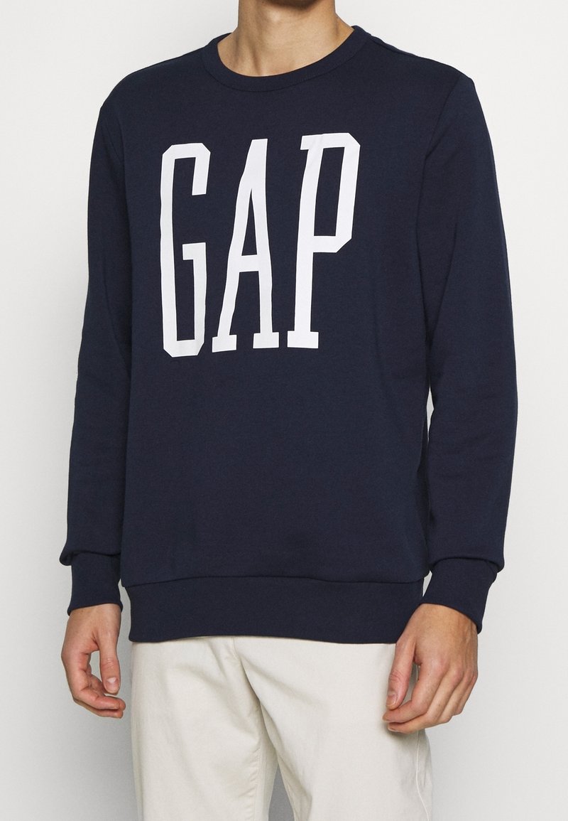Felpa blu navy con collo a giro, polsini e orlo a costine. Presenta un grande logo bianco "GAP" sul davanti. Tessuto morbido.