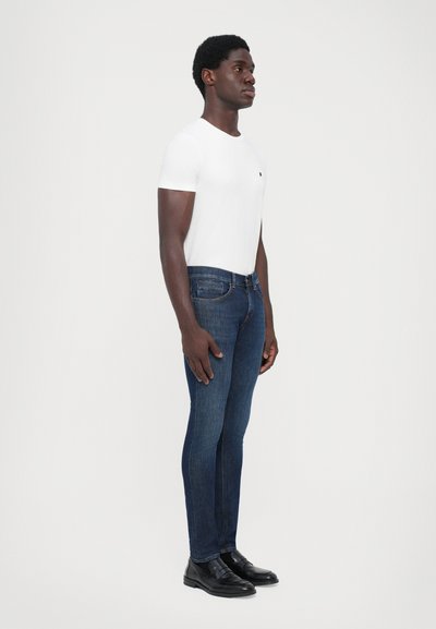 Jeans in denim blu scuro con una vestibilità slim, abbinati a una t-shirt bianca semplice. Scarpe formali nere completano l'outfit. Il modello è in piedi di lato.