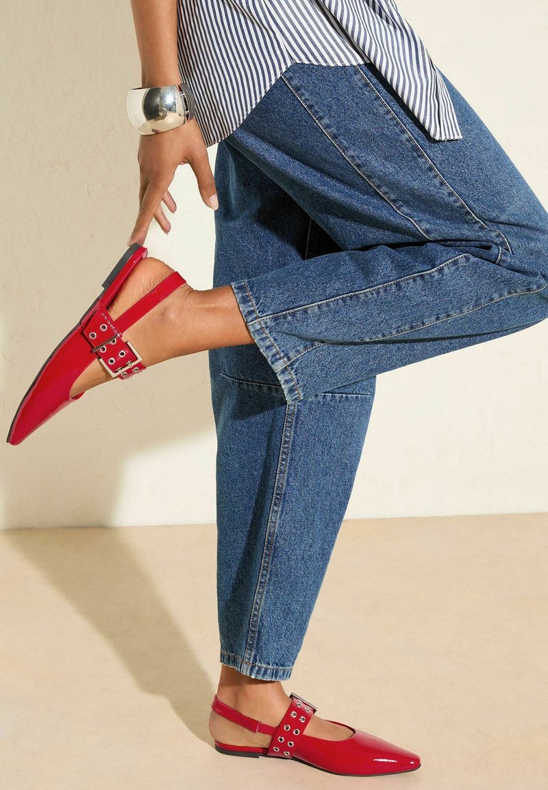Scarpa slingback in pelle verniciata rossa con punta quadrata, fibbia argentata e dettagli intagliati. Indossata con jeans in denim blu.