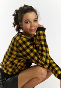 Maglione checkered giallo e nero con maniche lunghe e collo rotondo, abbinato a una gonna di pelle nera, mostrato in posizione accovacciata.