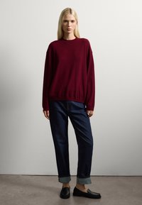 Femme blonde debout, portant un pull maroon ample, un jean bleu foncé retroussé et des mocassins noirs devant un mur uni.