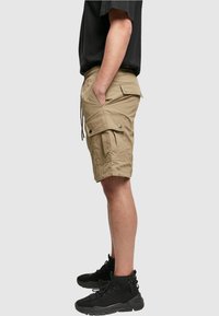 Beige cargo shorts, joissa on sivutaskut ja nepparit, joustava vyötärö nauhalla, ja kevyt kangas. Yhdistetty mustiin saappaisiin.