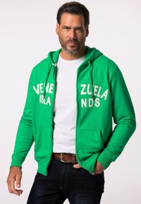 Sudadera verde con cremallera y texto blanco que dice "ISLAS VENEZOLANAS", con un bolsillo frontal, capucha con cordón y puños acanalados. Llevada sobre una camiseta blanca.
