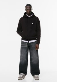 Felpa nera con cappuccio e lacci, logo bianco sul petto, abbinata a jeans grigi sbiaditi e larghi e sneakers grigie con dettagli bianchi.