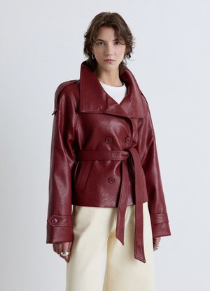 Woman wearing a belted burgundy leather jacket over a white top and cream pants, standing against a plain light background.