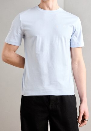 Man draagt een effen lichtblauw T-shirt met korte mouwen en zwarte broek, staat met linkerarm achter de rug en rechterarm ontspannen.