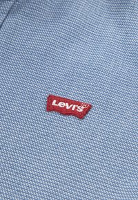 Lichtblauwe stof met een gestructureerde weving; heeft een rode geborduurde Levi's logo patch in de onderste hoek.