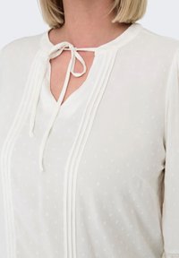 White blouse featuring a V-neck with a tie closure, pleated design, textured fabric with small dots, and long sleeves.