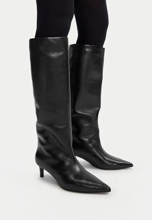 Botas de cuero negras hasta la rodilla con punta puntiaguda y tacones bajos, usadas con medias negras sobre un fondo liso.