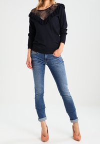 Marineblauer Pullover mit Spitzenpaneel und Rüschendetails, kombiniert mit hellblauen Jeans. Das Model trägt rosafarbene Absätze, die eine figurbetonte Silhouette zur Geltung bringen.