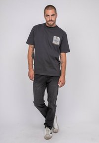 T-shirt en coton noir avec col rond, comportant une poche poitrine avec un imprimé graphique blanc. Associé à un jean foncé et des baskets blanches.