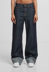 Plačios džinsinės kelnės tamsiu skalbimu, su kontrastingomis oranžinėmis siūlėmis ir laisvu kirpimu. Akcentai: sukrautas kraštas ir šoninės kišenės.