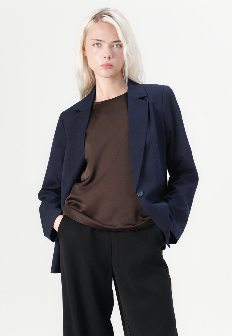 Blazer bleu marine avec une coupe ajustée, fermeture à un bouton et revers structurés, associé à un top en satin marron et un pantalon large noir.