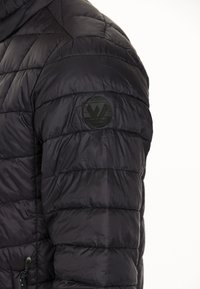 Sort pufferjakke med quiltet tekstur og glans. Har et rundt logo-patch på skulderen og en lynlåslomme på siden.