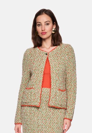 Woman wearing a multicolored textured jacket and matching skirt, with a bright orange top and large gold hoop earrings.