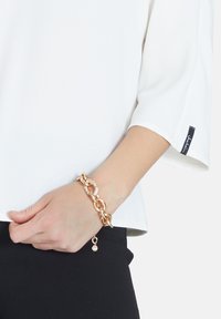 Weißes Blusenshirt mit Dreiviertelärmeln, kombiniert mit einem goldenen Armreif, der über runde Glieder und funkelnde Akzente verfügt. Schwarze Hose sichtbar.