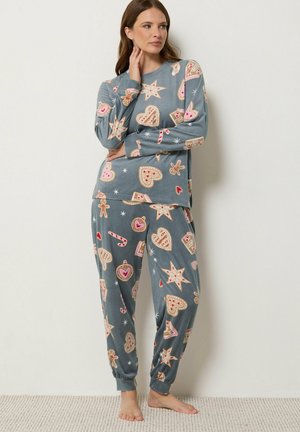 Femme en pyjama festif bleu-gris décoré de cœurs, d'étoiles, de pain d'épices, de cannes en sucre d'orge et de biscuits de Noël, debout pieds nus