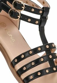 Sandalia negra de tiras hecha de material sintético, con tachuelas y hebillas en tono dorado, y plantilla acolchada beige.