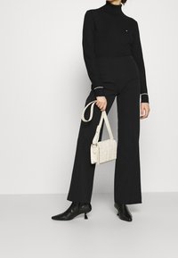 Femme portant un pantalon taille haute noir, un col roulé noir, des bottines à talons aiguilles noires, tenant un petit sac en toile tissé blanc par la anse.